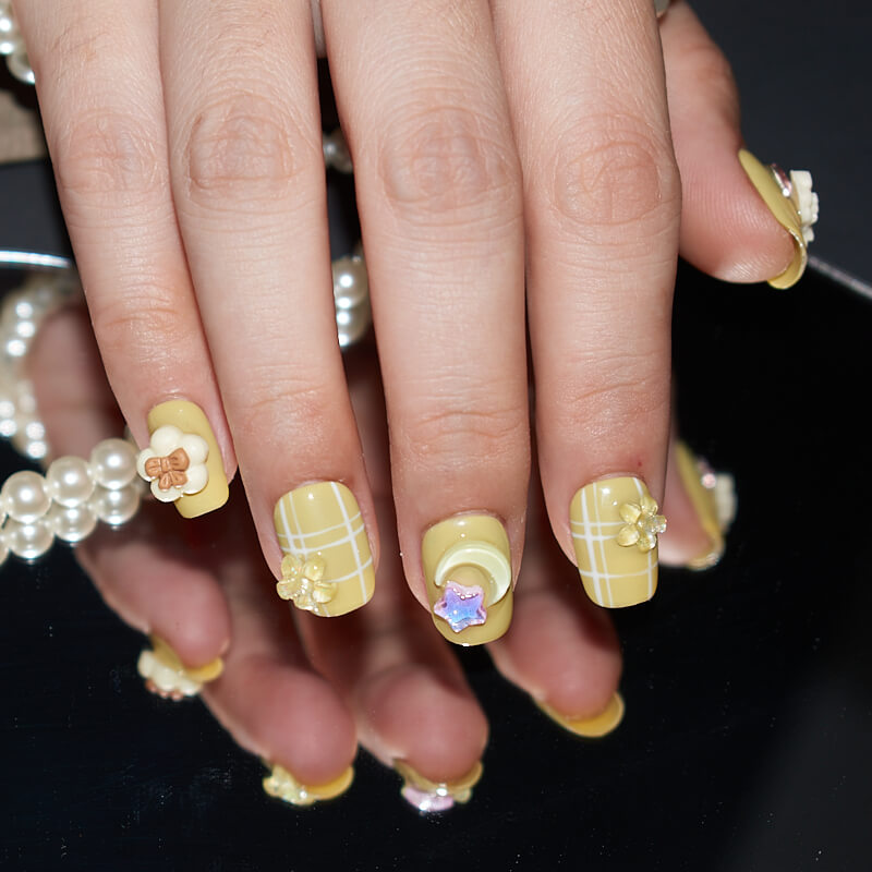 Hands with short, square-shaped yellow press-on nails featuring white grid patterns and 3D flower and star accents.