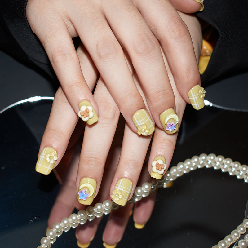 Hands with short, square press-on nails in yellow with white grid patterns and 3D flower and star accents.