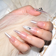 Hand delicately holding sheer fabric, highlighting elegant stiletto press-on nails with a pink ombre design and 3D butterfly and fairy accents."