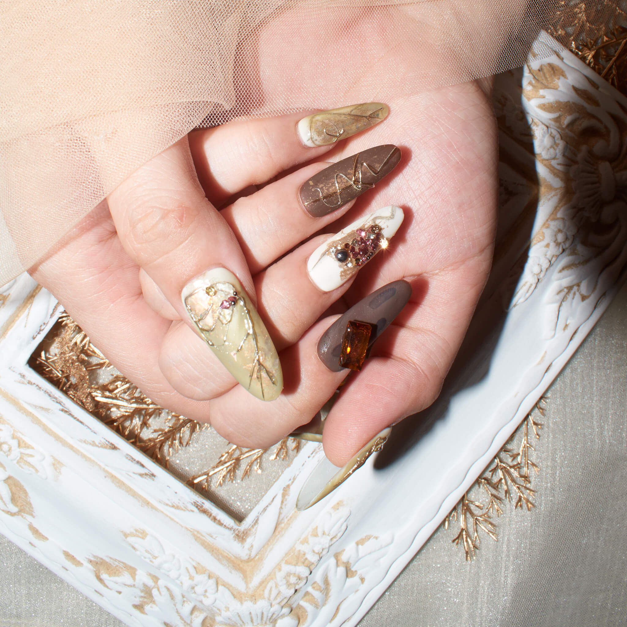 Hands with long, almond-shaped press-on nails in earthy tones with marble patterns, gold accents, and 3D embellishments.