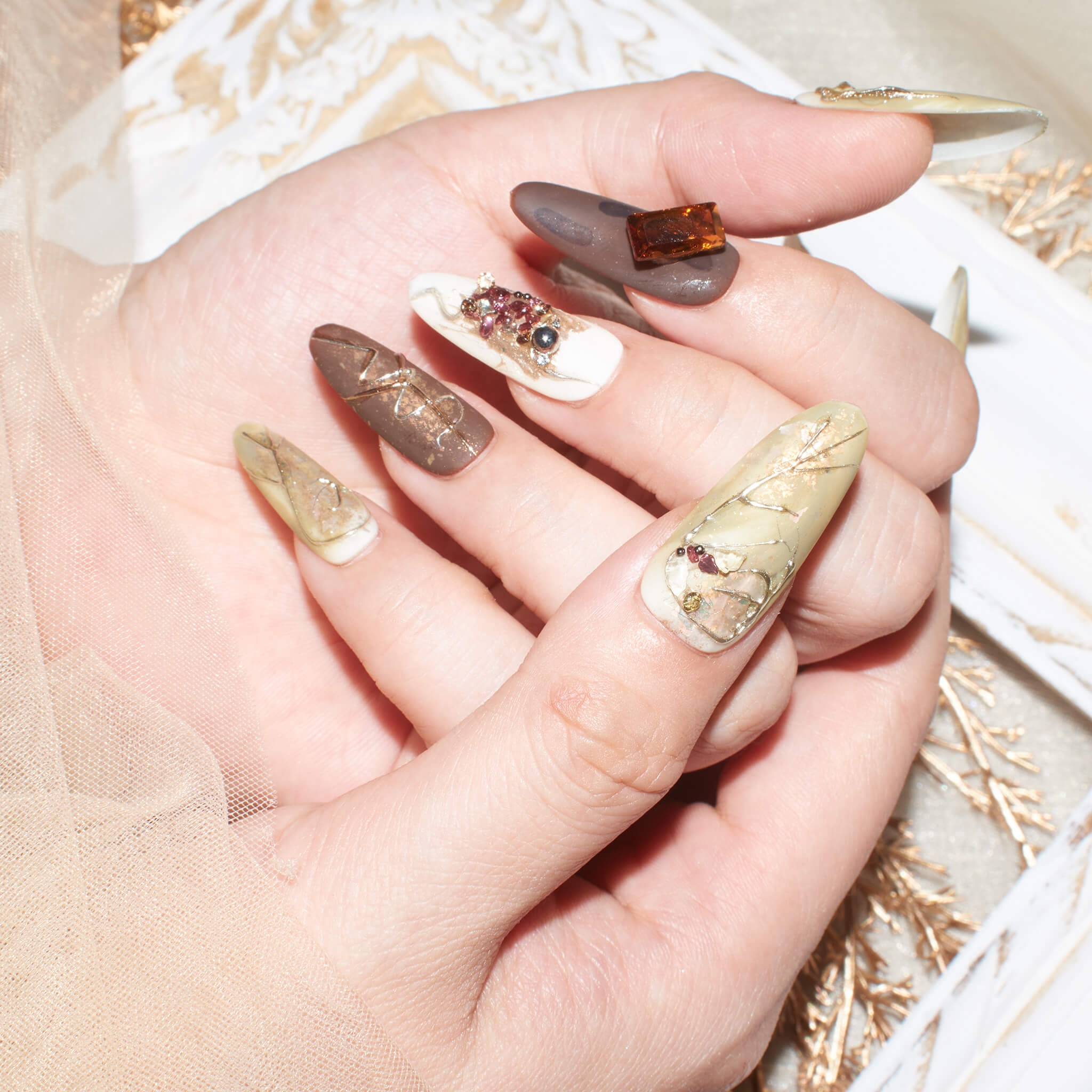 Hands with long, almond-shaped press-on nails in earthy tones of beige, brown, and taupe, featuring marble patterns, gold accents, and 3D embellishments such as jewels and geometric designs.