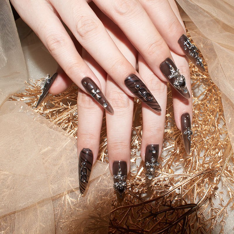 Hands with long, stiletto-shaped press-on nails in dark, smoky hues, adorned with silver accents, pearls, chains, and cross embellishments.